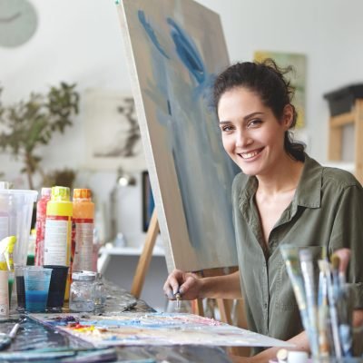Pleasant-looking smiling female in casual clothes, drawing abstract picture on easel while using colorful watercolors, holding brush in hand. Gorgeous female painter involved in creative process